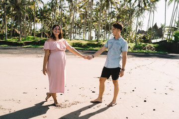 Wall Mural - Happy pregnant couple holding hands while standing on sandy coast