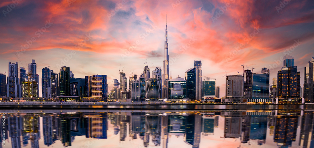 Stunning panoramic view of the Dubai skyline at sunset with buildings and skyscrapers reflected on a silky smooth water flowing in the foreground. Dubai, United Arab Emirates. - obrazy, fototapety, plakaty 