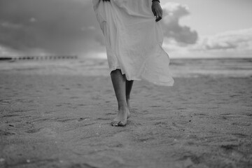 Poster - A greyscale shot of a beautiful lady's legs with a long white dress walking on the beach