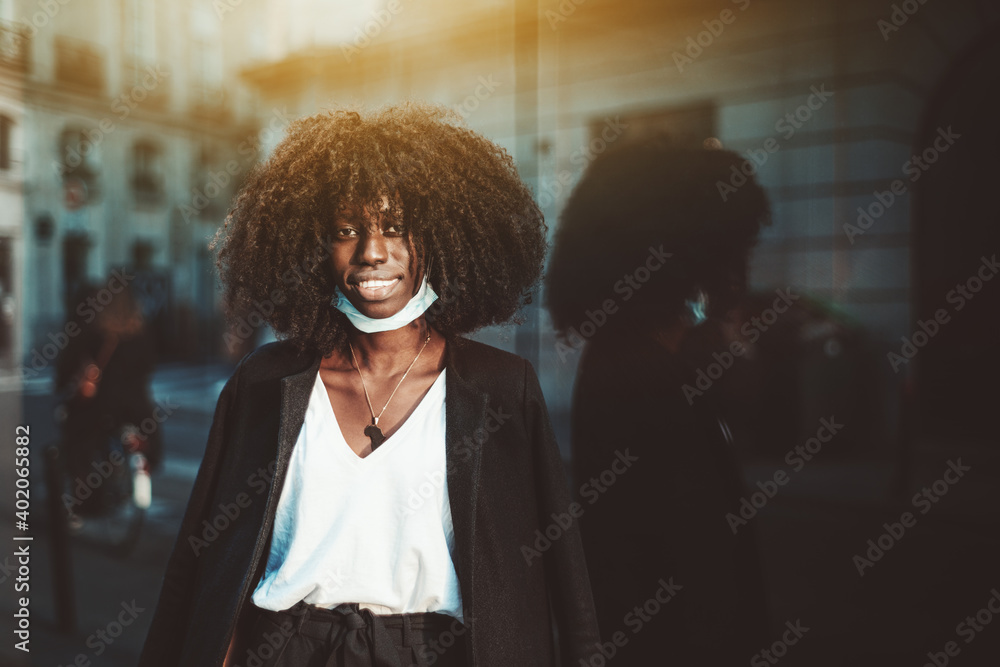 Portrait of a young beautiful cheerful black woman in a lowered virus protective mask, with a coat thrown over her shoulders, she's on the street leaning against a glass wall facade reflecting her - obrazy, fototapety, plakaty 