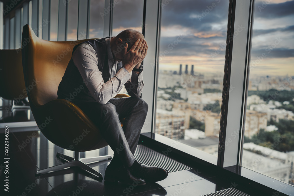 A tired broken man entrepreneur is worrying about the crisis and bankruptcy of the partner company while sitting on an armchair next to the window of a luxurious office skyscraper; a dark sky behind - obrazy, fototapety, plakaty 
