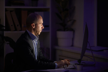 Side view shot of serious Caucasian man sitting alone in office room working on computer late at night, copy space