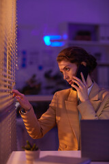 Vertical medium portrait shot of modern young businesswoman spending evening in office talking on phone with client