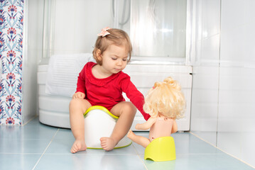 Beautiful smiling little baby sitting on potty in bathroom. Cute adorable funny child girl using chamber pot together with her baby doll. Toilet training concept. Toddler learning to use the Toilet.