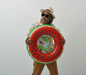 Happy elderly traveler asian man wearing summer shirt, straw hat and sunglasses holding inflatable ring standing over white wall background, Funny face expression, Business summer holiday concept