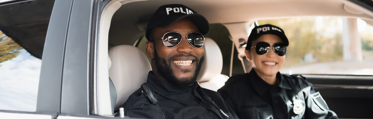 Poster - happy multicultural police officers looking at camera in patrol car on blurred background, banner.