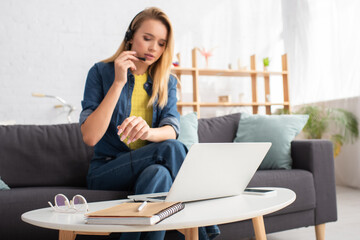  woman in headset sitting near coffee table with devices, notebook and eyeglasses at home on blurred background