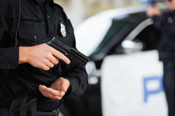 Cropped view of policeman holding firearm near car on blurred background outdoors.