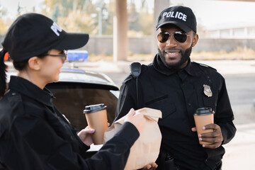 Poster - happy policewoman with paper cup giving package to african american colleague near patrol car on blurred background outdoors.