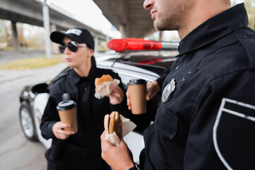 Wall Mural - Policeman holding burger and coffee to go near colleague and car on blurred background on urban street.