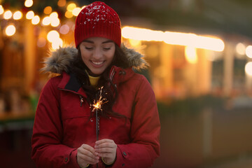 Wall Mural - Happy young woman with sparkler at winter fair in evening. Christmas celebration