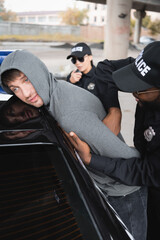 Wall Mural - african american policeman frisking sad hooded offender leaning on car on blurred background outdoors.