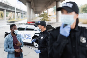 Wall Mural - African american victim in medical mask standing near police officer and policewoman on blurred foreground on urban street.