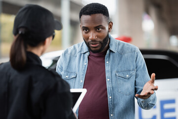 Wall Mural - back view of young policewoman with digital tablet near irritated african american victim on blurred foreground outdoors.