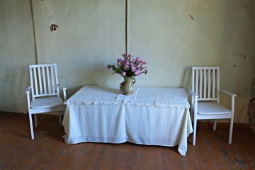 Bouquet of flowers on a table covered with a white tablecloth and two chairs in a room of an old house