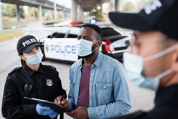 Wall Mural - African american victim in medical mask holding pen near police officer with clipboard and policeman on blurred foreground.