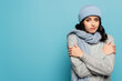 © LIGHTFIELD STUDIOS - brunette woman in hat and scarf hugging herself while looking at camera isolated on blue
