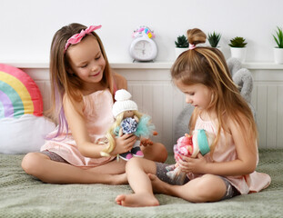 Little kid girls barefooted sisters in home clothing and headband with bow play with her favorite dolls in bed at home. Happy childhood, cheerful lifestyle, games, comfortable pastime, hobby concept