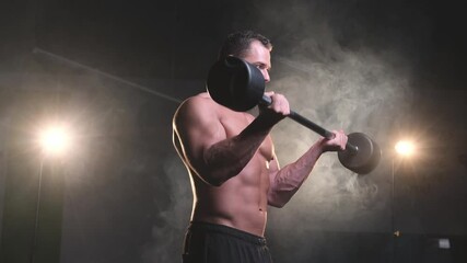 Wall Mural - Sexy caucasian athlete with naked torso and muscular build doing physical exercise lifting a dumbell in dark and atmospheric room with smoke. Wellness and healthy lifestyle of a sportsman in solitude.