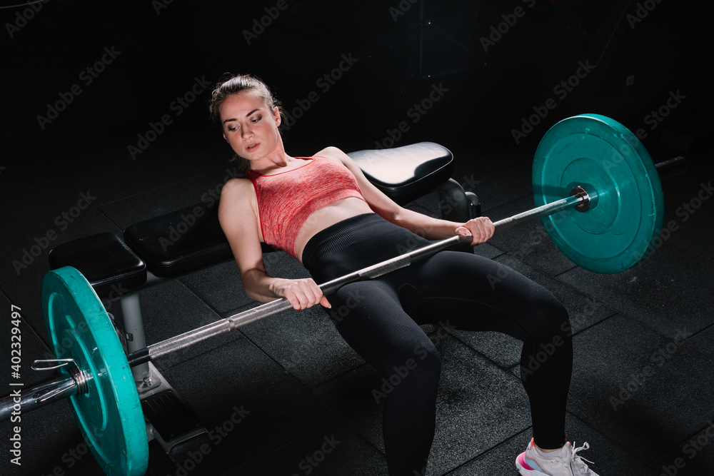 Young beautiful woman doing hip thrust. She poses with barbells in the gym. - obrazy, fototapety, plakaty 