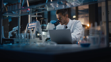 Medical Development Laboratory: Portrait of Male Scientist Works on Microscope, Using Laptop, Enters Data for Analysis. Pharmaceutical Lab for Medicine, Biotechnology, Drugs, Research. Evening Work