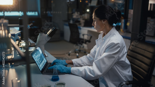 Medical Development Laboratory: Female Scientist Works on Laptop, Enters Data for Further Analysis. Pharmaceutical Lab for Medicine, Biotechnology, Bio Chemistry, Drugs, Vaccine Research. Evening Work
