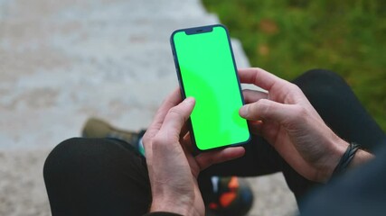 Outdoors over the shoulder shot of man hands use phone with vertical green screen outdoors. Sitting. Touchscreen finger internet cellphone scrolling. Close up. Slow motion