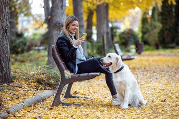 Sticker - Attractive young woman sending messages with her smart phone while sitting on bench with her lovely dog in the park in autumn.