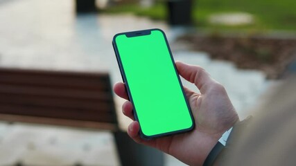 Outdoors young hands man holding use phone with vertical green screen in park background bench. Internet finger gadget device. Close up. Slow motion