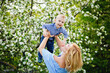 © Elena  - A woman plays with a child on the street in a blooming spring garden. Mother lifts her son in her arms up against the background of apple trees with flowers.