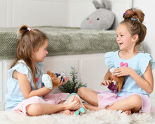 Happy Laughing Kid Girls Sisters In Blue Dresses Play Funny Situations With Their Dolls On Soft Carpet Rug At Home. Happy Childhood, Cheerful Lifestyle, Games, Comfortable Pastime, Hobby Concept