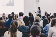© kasto - Business and entrepreneurship symposium. Female speaker giving a talk at business meeting. Audience in conference hall. Rear view of unrecognized participant in audience. Copy space on whitescreen.