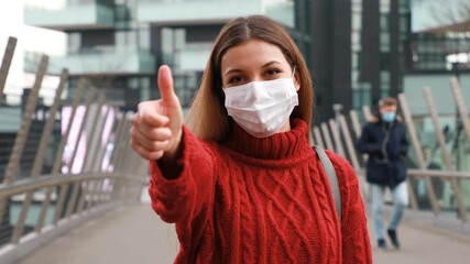 Wall Mural - COVID-19 Optimistic young woman wearing protective mask avoiding Coronavirus disease 2019 showing thumbs up in city street