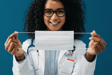 Wall Mural - Close up of a female doctor holding a mask in her hands. Woman in medical uniform looking at camera at blue background.