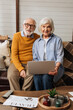 © LIGHTFIELD STUDIOS - happy elderly couple with laptop looking at camera on couch on blurred foreground in living room
