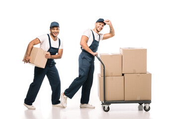 Wall Mural - full length of positive multicultural movers in uniform and caps walking near hand truck with carton boxes on white