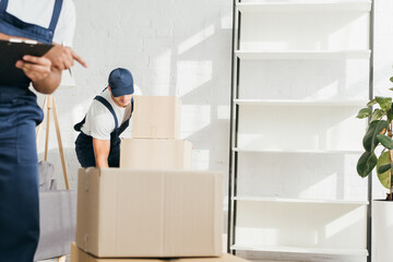 Wall Mural - young mover in cap carrying boxes near indian coworker on blurred background