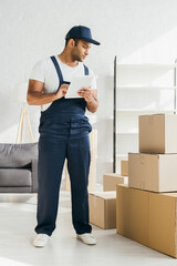 Wall Mural - full length of indian worker in uniform holding digital tablet and looking at boxes in apartment