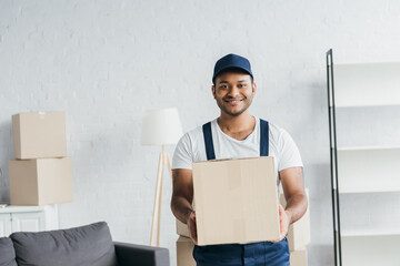 Wall Mural - young cheerful indian courier in cap and uniform holding box in apartment