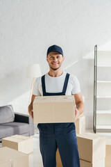 Wall Mural - tattooed and smiling mover in cap holding carton box and looking away in apartment