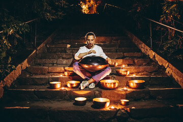 silhouette of a man playing handpan surrounded by Tibetan singing bowls and lighted candles sitting on the steps of the night city. Noise effect on the film