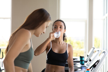 young hispanic woman and friend in sportswear running on treadmill at gym and holding bottle of water