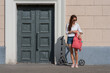 © byswat - girl in summer in city straightens her backpack bag, with a scooter, casual clothes denim shorts, a white shirt and a pink backpack. Background wall and old wooden door entrance to the building.