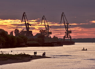 Crimson sunset over the river Ob. Silhouettes of fishermen on the shore and cranes on the horizon