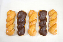 Top View Flat Lay Of Row Of Alternating Glazed Twist Donut And Chocolate Covered Glazed Twist On Parchment Paper.
