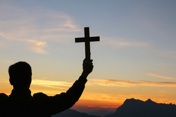 Silhouette human praying to the GOD while holding a crucifix symbol with bright sunbeam on mountain at sunset time