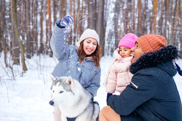 Wall Mural - afro man with his caucasian wife having fun with a beautiful daughter playing husky in snowy park