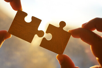 two hands trying to connect puzzle piece. sunset sky background. Jigsaw wooden puzzles against sun rays. one part of the whole. symbol of association and connection. business strategy. copy space