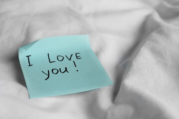 Sticky note with phrase I love you! on bed, closeup. Space for text
