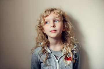 Wall Mural - Pretty beautiful pensive Caucasian blonde girl with long hair on light neutral beige background wall. Child kid thinking dreaming about future. Happy authentic childhood lifestyles.